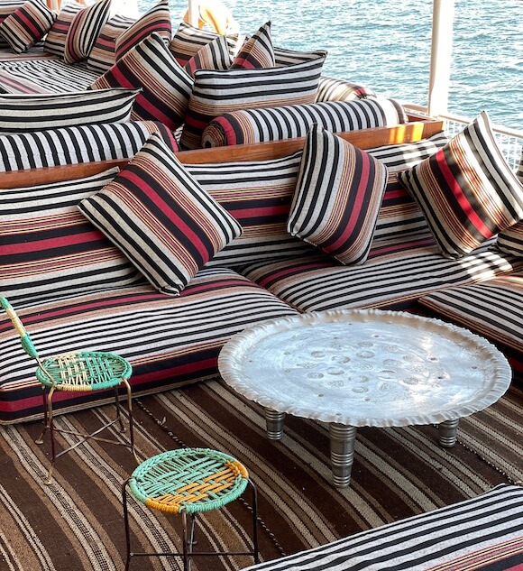 Lounging area on a boat with striped pillows and metal tray with green woven seats.